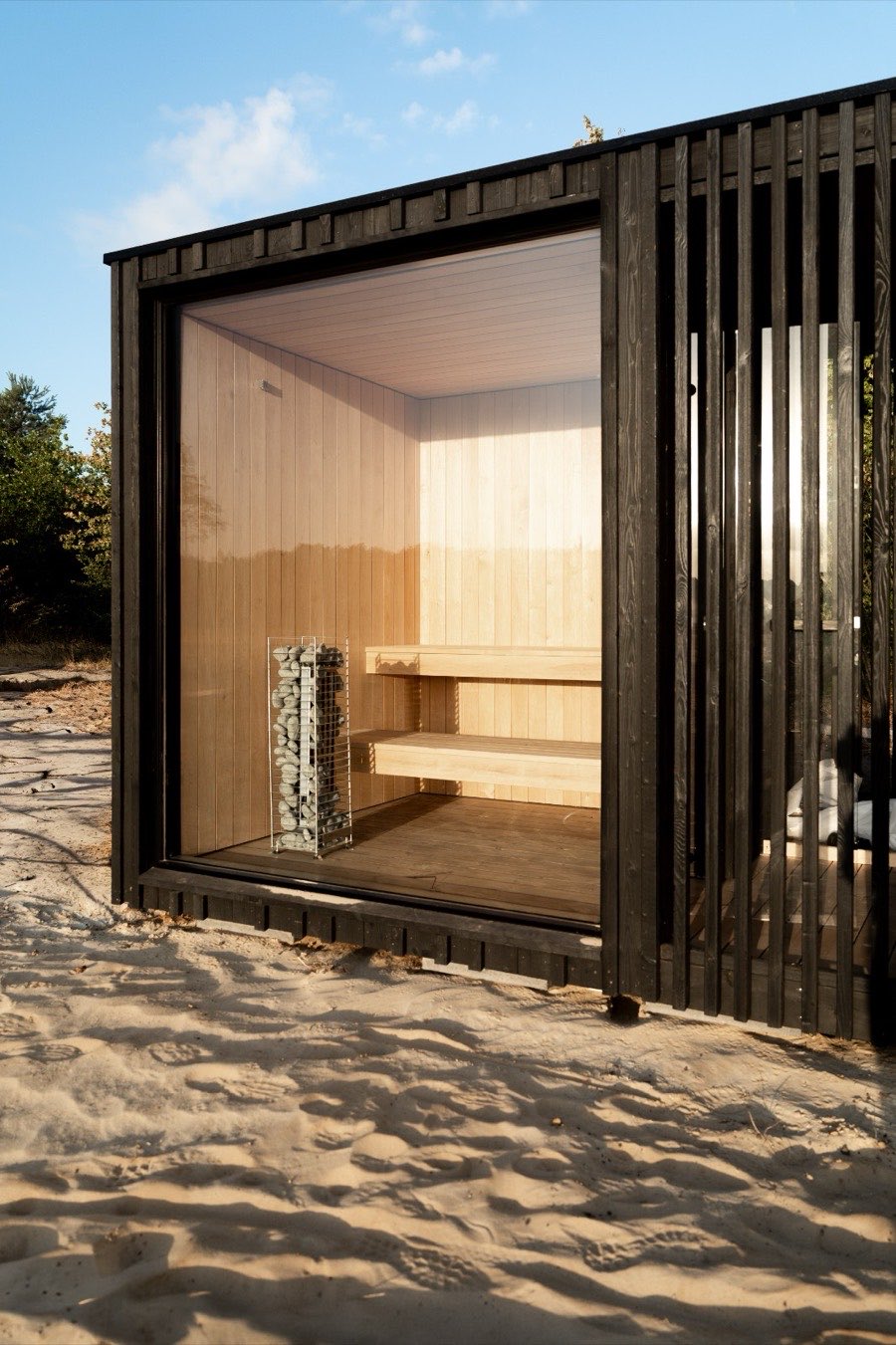 De sauna in het zand, met zwarte houten lamellen en een glazen front waarachter de banken en de kachel staan